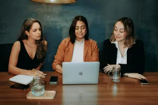 Three women engaged in a focused business meeting at a modern office setting with laptops and notebooks.