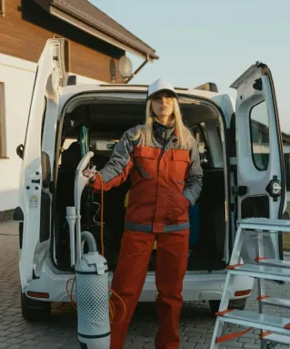 A cleaner in uniform stands beside a van with cleaning tools outside a modern house at sunset.