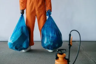 Sanitation worker in orange overalls holding blue trash bags indoors for waste management.
