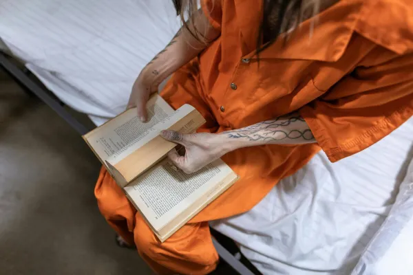 Female prisoner reading a book while sitting on a bed, wearing orange uniform.