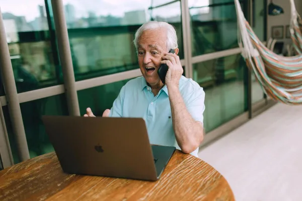 Senior adult happily chatting on phone and laptop, sitting indoors at a table.