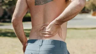 Shirtless man holding his lower back outdoors indicating pain or discomfort.
