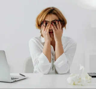 A stressed woman in eyeglasses and white shirt sits at her desk, overwhelmed and exhausted.