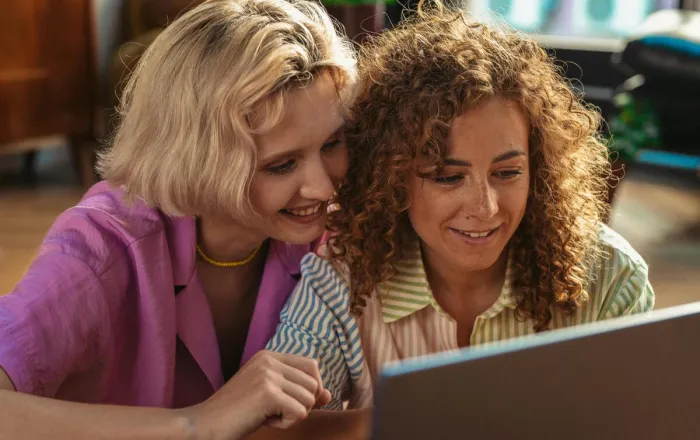 Two women smiling and working together on a laptop in a cozy indoor setting, fostering teamwork and connection.