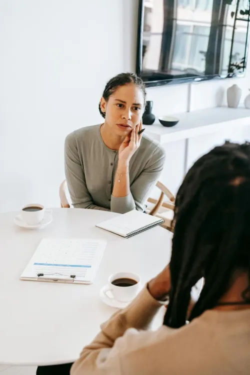 Serious ethnic female consultant listening to black male client problems while sitting at round table together in light psychology center