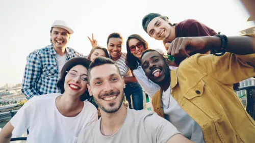 A diverse group of friends smiles while taking a selfie outdoors during the day.