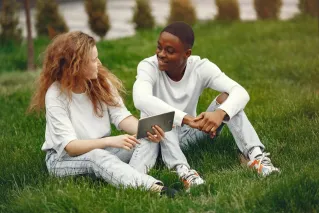 An interracial couple sitting on grass, smiling and using a tablet together.