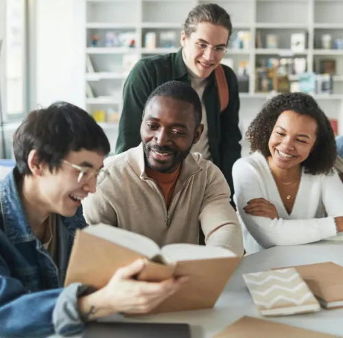A group of diverse students smiling and studying together indoors in a library setting.