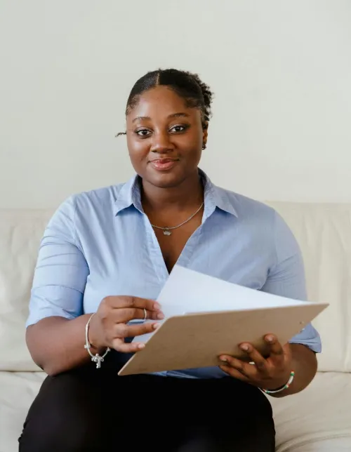 A confident woman in blue shirt sitting on a white sofa holding a clipboard, smiling.