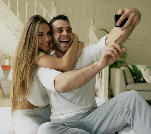 Smiling couple sitting on the bed, capturing a joyful selfie indoors.