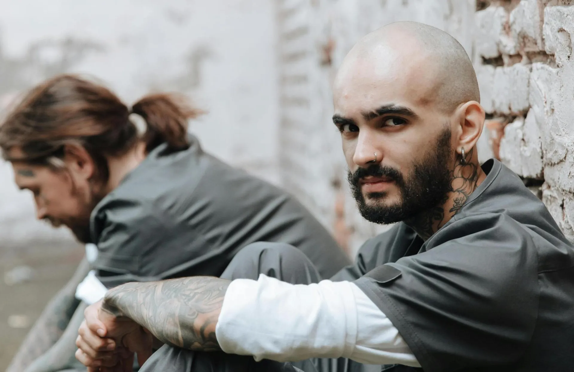 Two inmates with tattoos sitting against a brick wall wearing prison uniforms. Intense expressions.