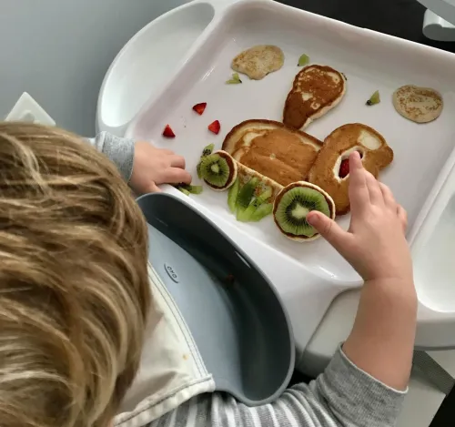 Child creating a playful breakfast art with pancakes and fruits on a high chair tray.