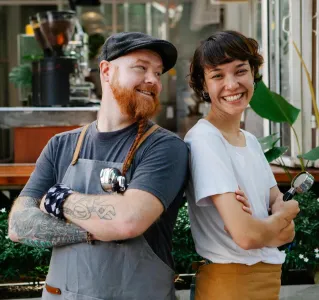Couple of smiling barista coworkers in casual outfit and aprons standing with portafilter near cafeteria in street near plants in daylight