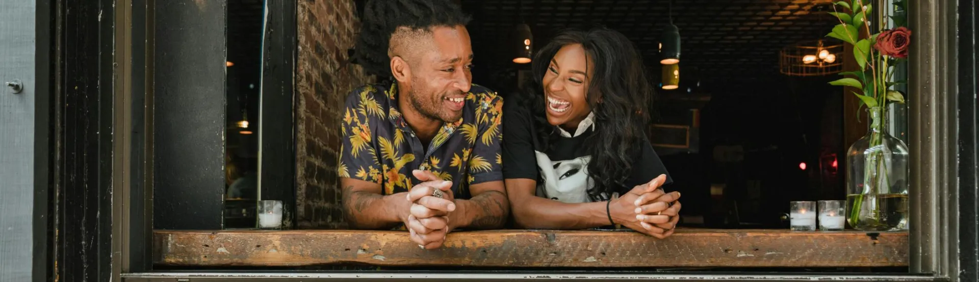 Two joyful business owners lean on a window sill with a 'Black Businesses Matter' sign visible.