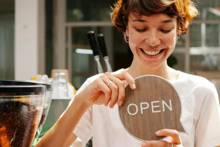 Happy barista holding open sign in a cozy café, ready to welcome customers.