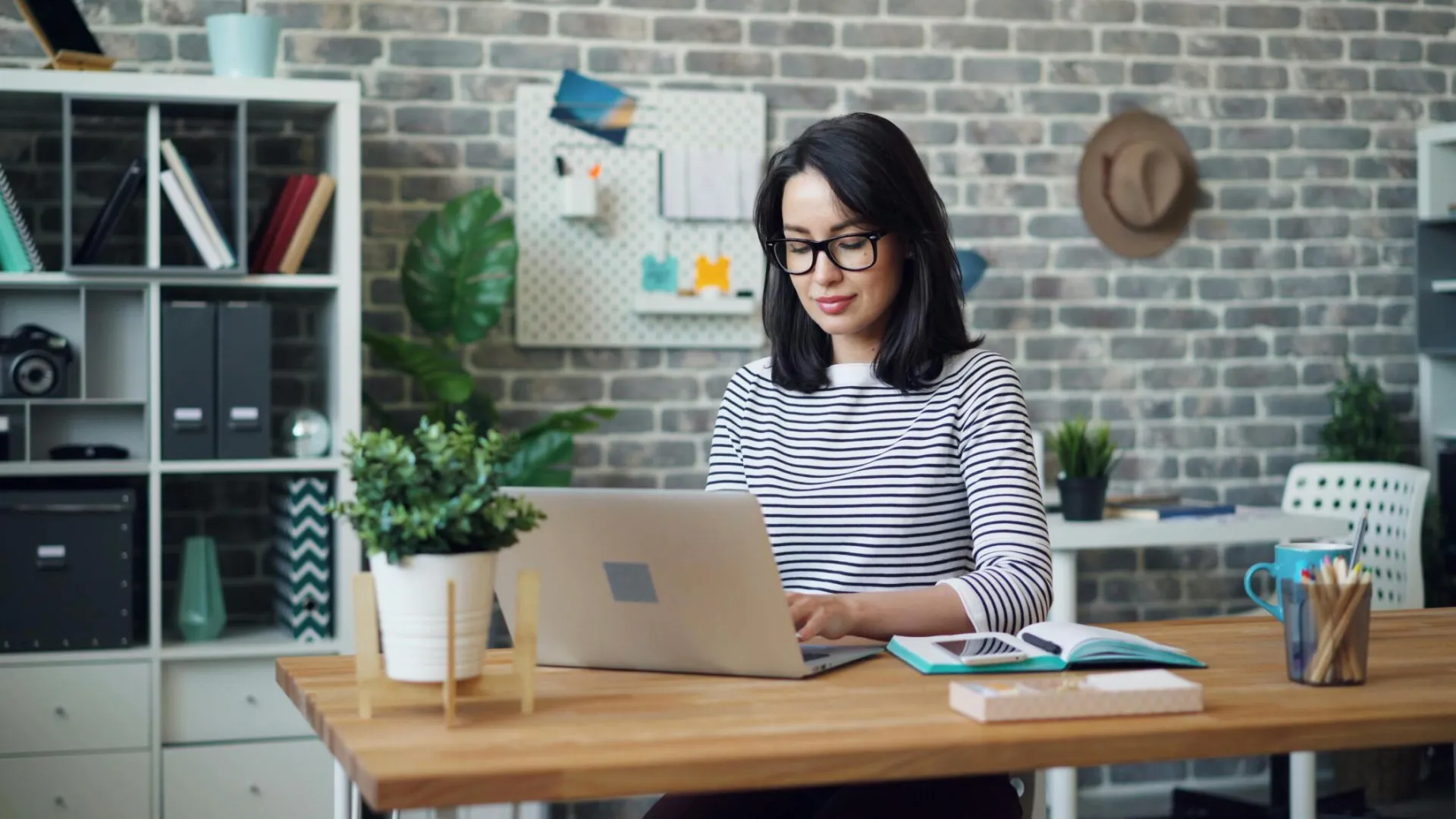 Young woman working on a laptop in a modern, stylish office setting with brick wall decor.