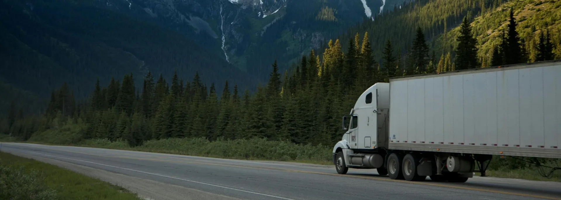 A semi-truck travels along a highway with snow-capped mountains in the background.