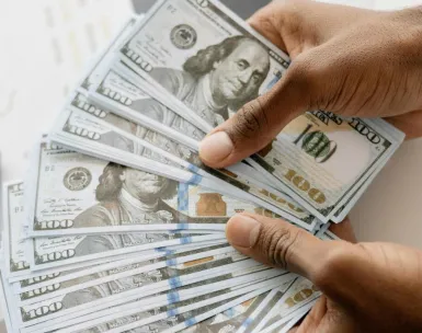 Close-up view of hands counting U.S. hundred dollar bills over a laptop background.