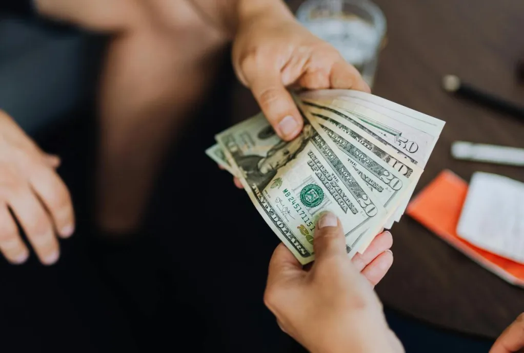 A close-up view of hands exchanging dollar bills indoors, symbolizing payment.