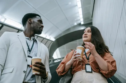 Two business professionals conversing casually on an escalator while holding coffee cups.