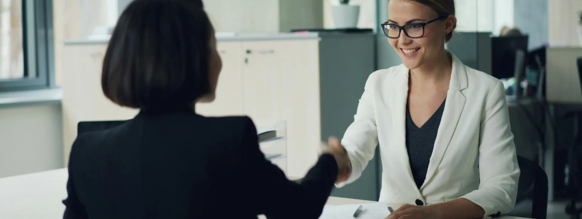 Two businesswomen shaking hands in a modern office setting, symbolizing partnership.