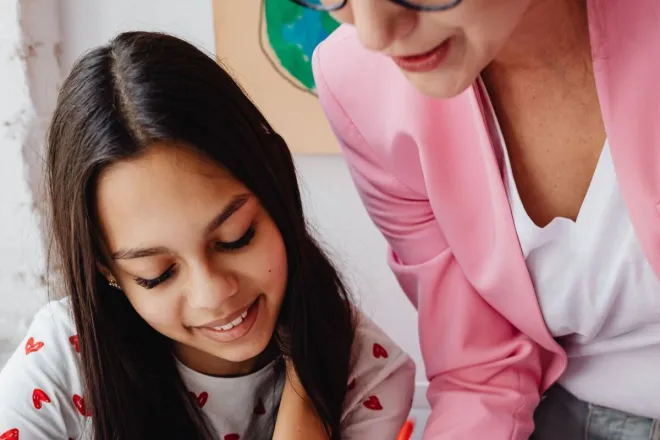 A teacher assists a student with a colorful school project at a table indoors.