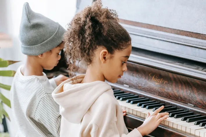 Side view of concentrated little African American girl with curly hair in casual clothes sitting on chair and playing piano with cute brother during weekend at home
