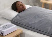 Woman lying in bed with a towel turban, applying facial skincare with eyes closed.