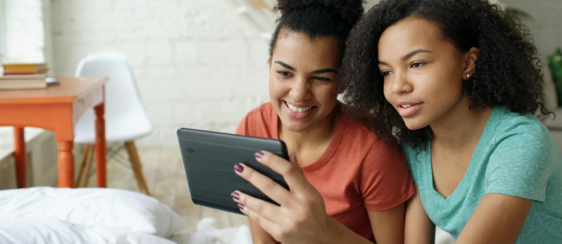 Two teenage girls using a tablet at home, smiling and enjoying their time together.