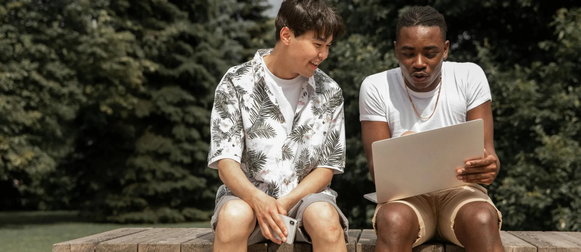 Smiling multiracial friends in stylish clothes sitting on wooden bench and discussing information from netbook