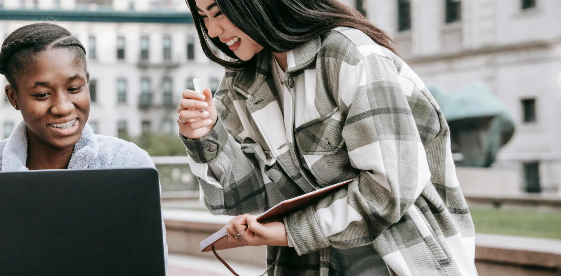 Young cheerful multiethnic female friends watching netbook while studying in town against buildings in daylight