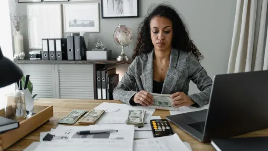 Focused woman counts cash at desk surrounded by office items, emphasizing finance and professionalism.