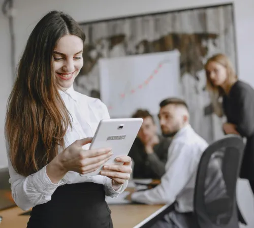 Young woman in a professional office setting browsing a tablet with colleagues in the background.