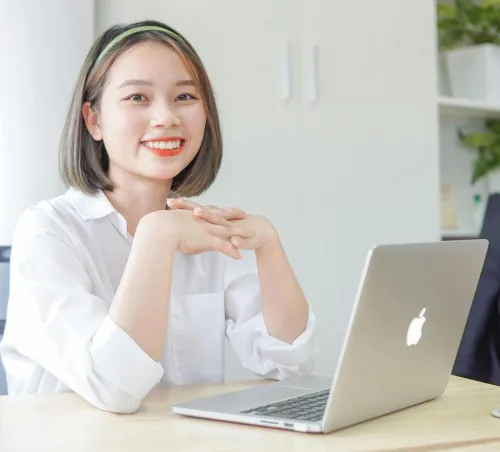 Woman sitting at desk with laptop, smiling confidently in a modern bright office setting.