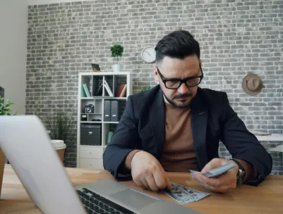 Businessman counting cash at a modern office desk with a laptop, showcasing finance management.
