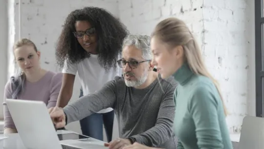 Bearded man wearing casual clothes sitting at table and working on laptop and explaining details of project to women while working
