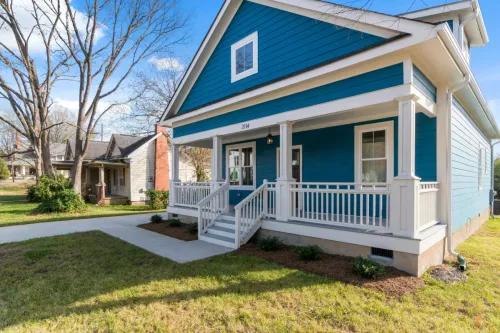 A cozy blue suburban house with a white front porch and lush surroundings on a clear day.
