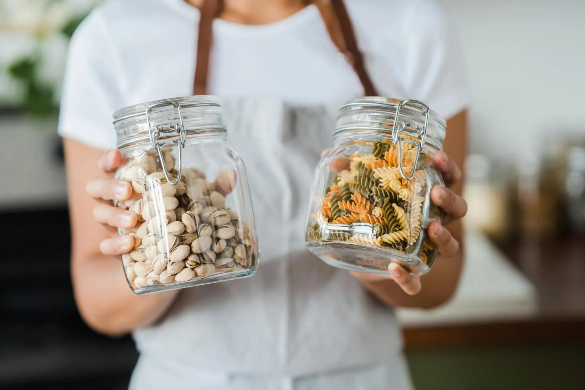 Hands holding glass jars filled with pistachios and colorful spiral pasta indoors.