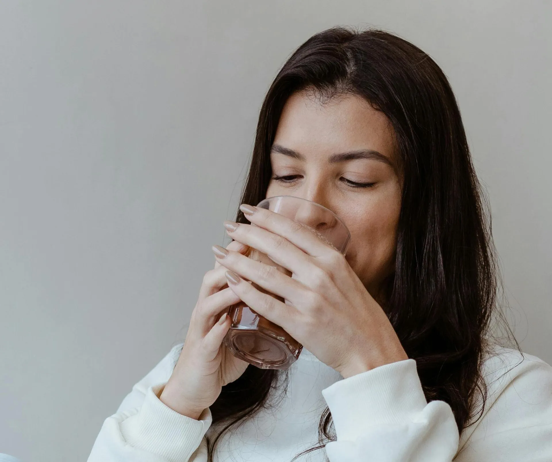 Brunette woman sitting indoors, sipping a drink with a relaxed expression and long hair.