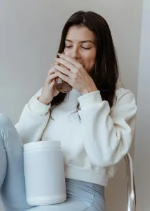 Brunette woman sitting indoors, sipping a drink with a relaxed expression and long hair.