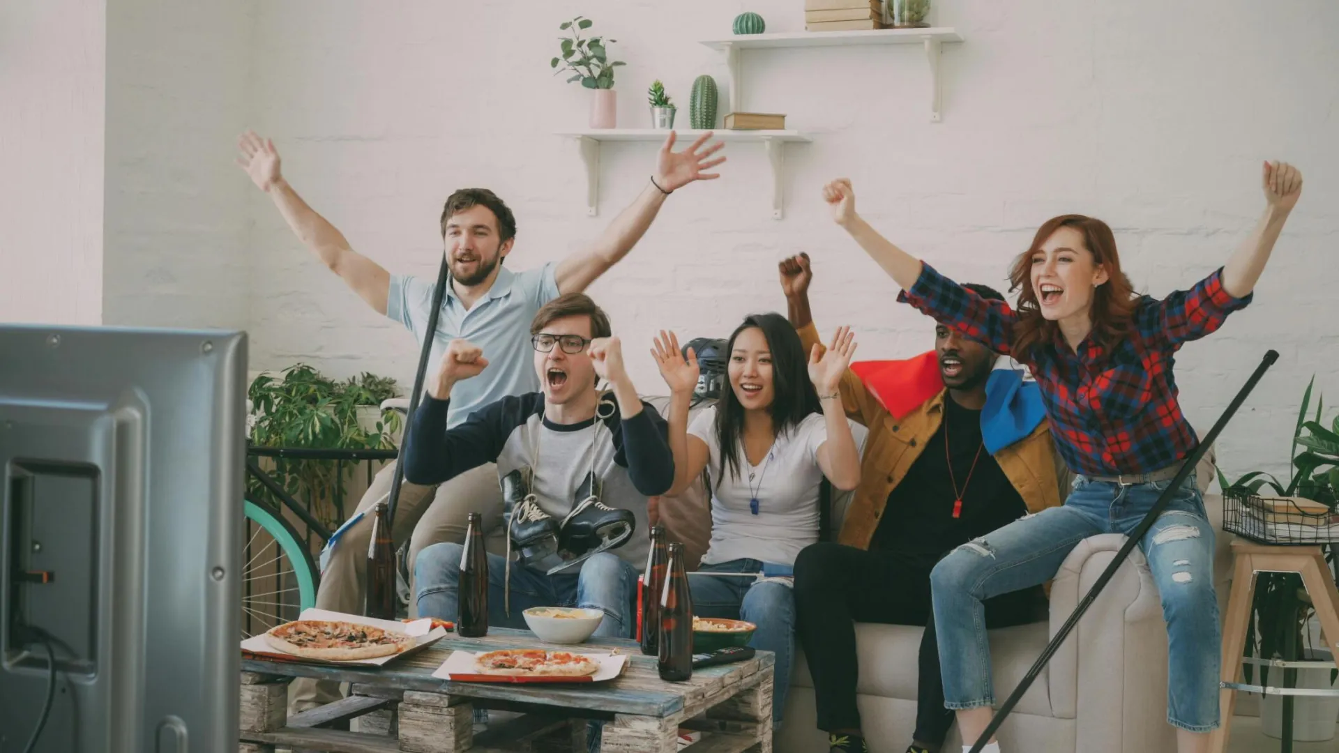 Group of friends cheering while watching sports on TV with pizza and drinks.