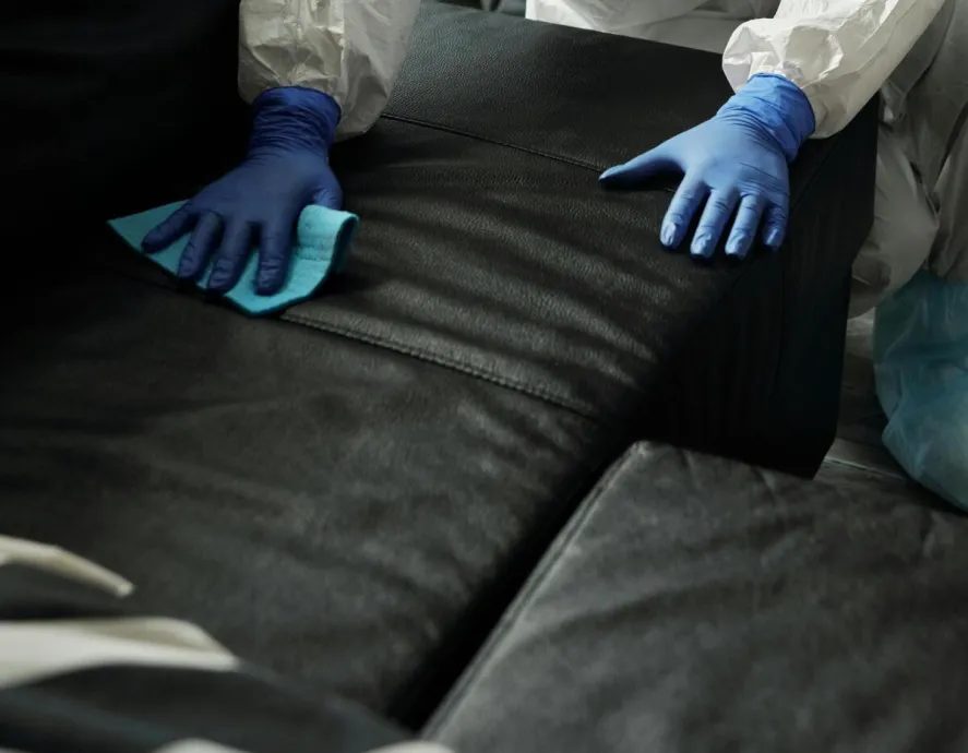 Person in full protective gear disinfecting a leather couch indoors with attention to hygiene and safety.