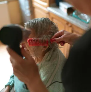 A caregiver assists a senior adult with hair drying and combing at home.
