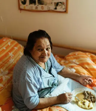 Senior woman sitting in bed with a meal, surrounded by family photos in a warm, cozy room.