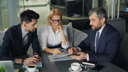 Three professionals engaged in a business discussion at an office table with coffee.