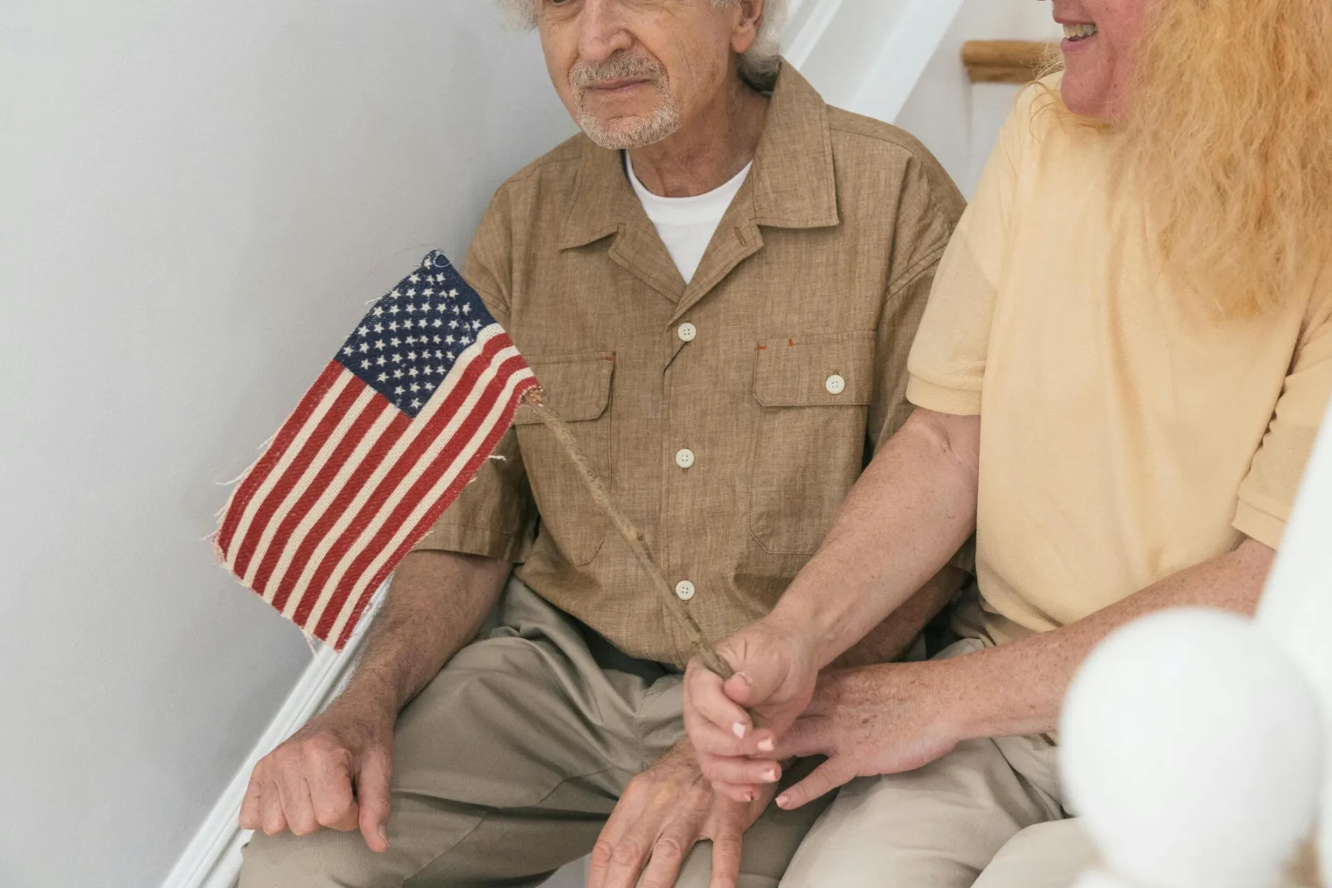 Senior couple sitting on steps indoors, holding a small United States flag together.