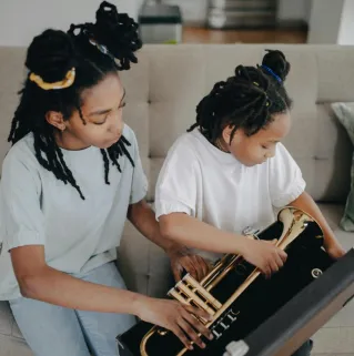 Two young girls with dreadlocks exploring a trumpet together indoors, fostering music interest.