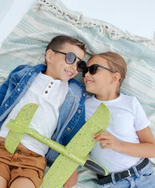 From above of cheerful child in sunglasses lying near smiling sister with plane on blanket and looking at each other