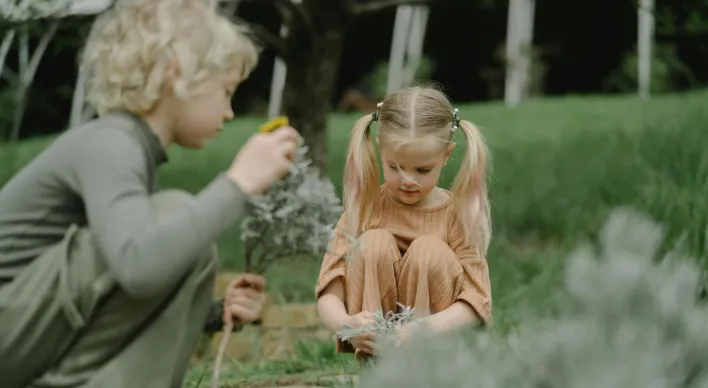 Two children engage in outdoor play and exploration, interacting with plants in a grassy garden.
