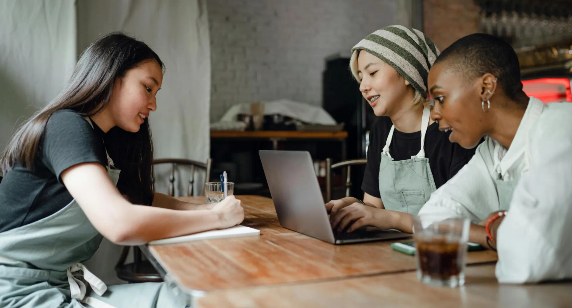 Positive smiling multiethnic female coworkers wearing casual clothes and aprons sitting at wooden table with modern laptop and taking notes while working on project together in creative workplace